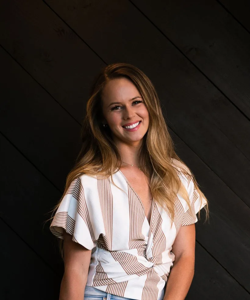 Tayler, Executive Director at Ela Construction, smiling in a striped wrap top against a dark wood-panelled wall.
