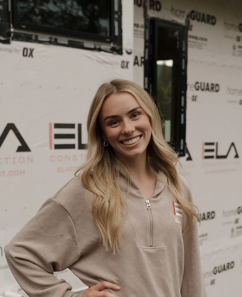 Tierra, Experience Coordinator at Ela Construction, in a tan Ela quarter-zip in front of house wrap printed with the Ela Construction logo.