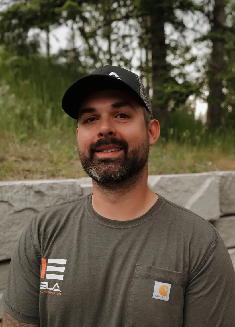 TJ, Operator at Ela Construction, in a black Ela cap and grey Ela T-shirt in front of a stone retaining wall.