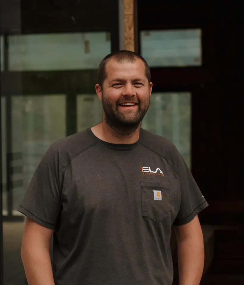 Tobin, Director of New Construction at Ela Construction, in a charcoal Ela Construction T-shirt in front of tall shop windows.
