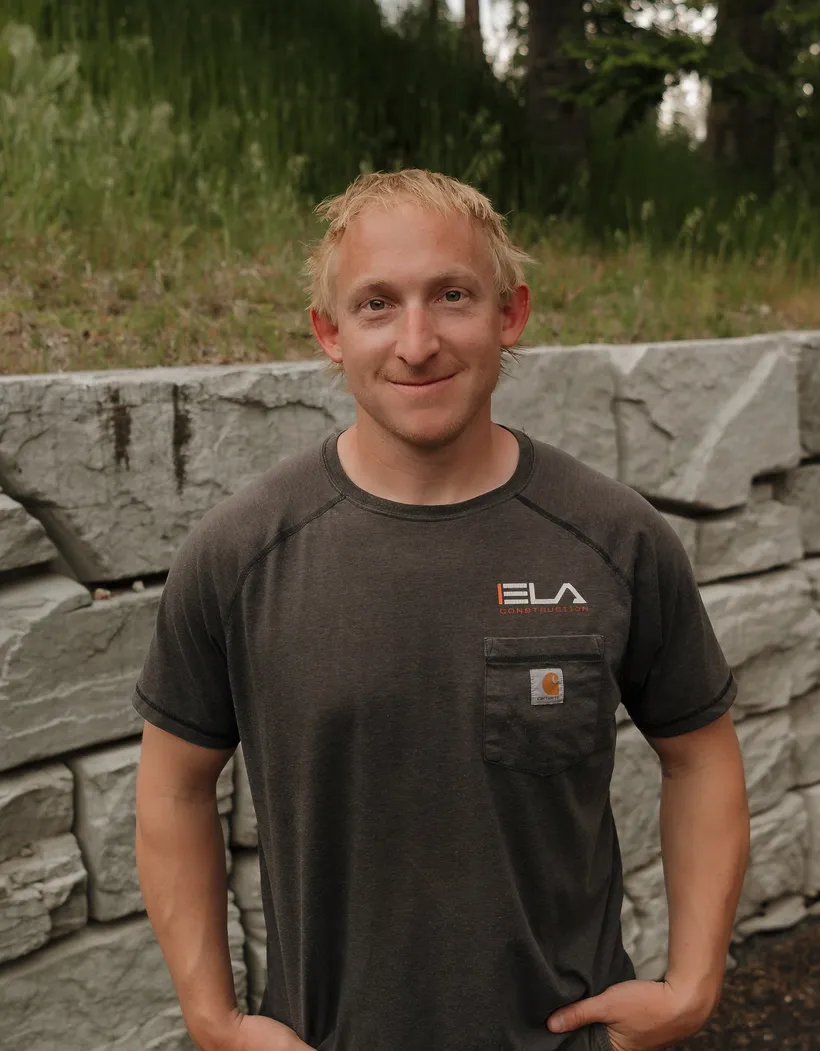 Warren, Owner of Ela Construction, smiling in a grey Ela Construction T-shirt in front of a stacked-stone retaining wall.